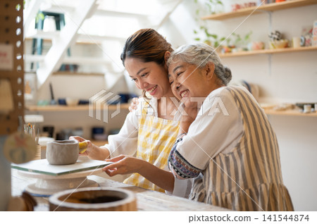 Senior Asian Mother Lovingly Leaning on Daughter's Shoulder at Pottery Workshop. Senior Asian Mother Lovingly Leaning on Daughter's Shoulder at Pottery Workshop. 141544874