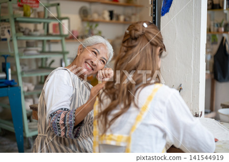 Senior Asian Mother Smiling Warmly at Adult Daughter During Craft Workshop in cozy studio Senior Asian Mother Smiling Warmly at Adult Daughter During Craft Workshop in cozy studio 141544919