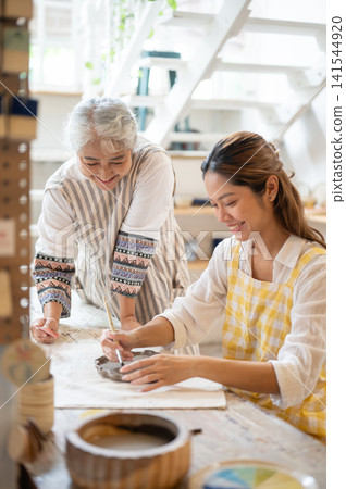 Senior Asian Instructor Watching Student Paint Ceramic Plate with Brush in pottery workshop. 141544920