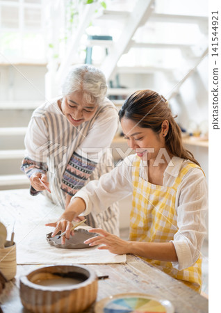 Elderly woman assists a younger woman with clay crafting during a warm and creative pottery workshop Elderly woman assists a younger woman with clay crafting during a warm and creative pottery workshop 141544921
