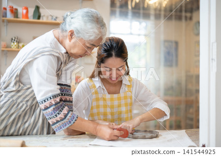Senior Asian Woman Helping Daughter Smooth Clay Plate with Scraper Tool in pottery workshop studio. Senior Asian Woman Helping Daughter Smooth Clay Plate with Scraper Tool in pottery workshop studio. 141544925
