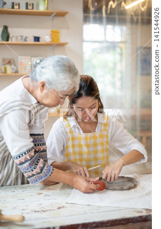 Senior Asian Woman Demonstrating Rib Tool Technique to Daughter in pottery workshop studio. Senior Asian Woman Demonstrating Rib Tool Technique to Daughter in pottery workshop studio. 141544926