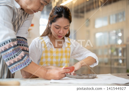 Asian Senior Instructor Overseeing Young Student Shaping Clay Plate in pottery workshop studio. Asian Senior Instructor Overseeing Young Student Shaping Clay Plate in pottery workshop studio. 141544927
