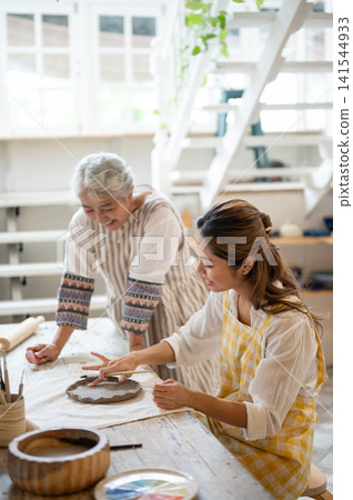 Smiling Asian Mother Watching Daughter Paint on Ceramic clay plate in pottery workshop studio. Smiling Asian Mother Watching Daughter Paint on Ceramic clay plate in pottery workshop studio. 141544933