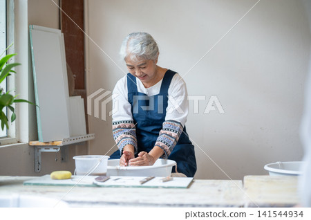 Senior Asian Woman Smiling as She Shapes Clay on Electric Potter's Wheel in pottery workshop studio. Senior Asian Woman Smiling as She Shapes Clay on Electric Potter's Wheel in pottery workshop studio. 141544934