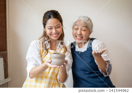 Asian Senior Woman Gesturing Admiration as Daughter Presents Clay Pot in pottery workshop studio. Asian Senior Woman Gesturing Admiration as Daughter Presents Clay Pot in pottery workshop studio. 141544962