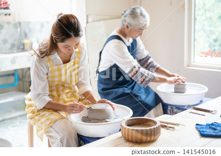 Asian Daughter Trimming Clay Vessel while Senior Mother Crafts on Wheel in pottery workshop studio. Asian Daughter Trimming Clay Vessel while Senior Mother Crafts on Wheel in pottery workshop studio. 141545016