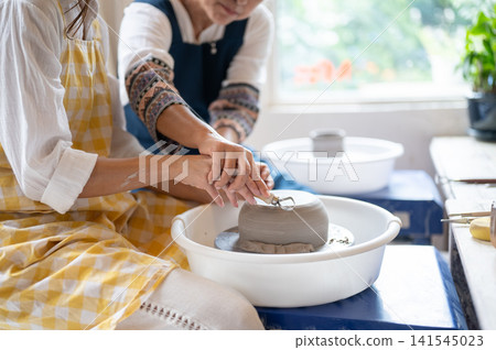 Close Up Senior Woman Hand Guiding Student Trimming Clay Bowl in pottery workshop studio. Close Up Senior Woman Hand Guiding Student Trimming Clay Bowl in pottery workshop studio. 141545023