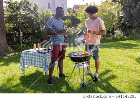 Diverse male friends grilling corn, burgers on charcoal grill in backyard holding drinks Diverse male friends grilling corn, burgers on charcoal grill in backyard holding drinks 141545295