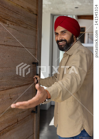 Mid adult indian man wearing red turban pulling door, extending hand in foyer, copy space Mid adult indian man wearing red turban pulling door, extending hand in foyer, copy space 141545314