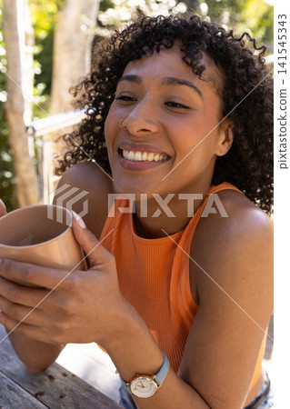 African american woman in orange tank top smiling while holding ceramic mug at patio table African american woman in orange tank top smiling while holding ceramic mug at patio table 141545343