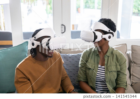 African american couple using vr headsets while sitting on couch by french doors interacting African american couple using vr headsets while sitting on couch by french doors interacting 141545344