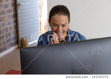 Woman wearing denim shirt typing at desk in home office with computer monitor, shuttered window Woman wearing denim shirt typing at desk in home office with computer monitor, shuttered window 141545372