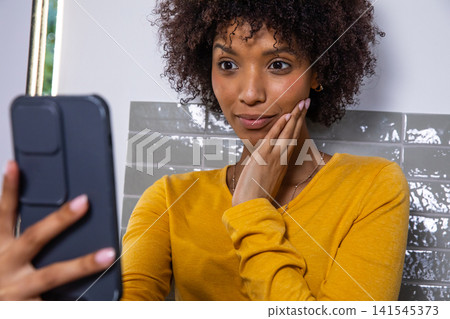 African american woman in yellow top holding smartphone near face checking skin in bathroom African american woman in yellow top holding smartphone near face checking skin in bathroom 141545373