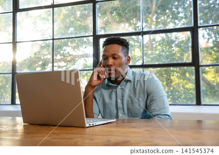 African american man sitting at wooden desk in home office gazing at laptop through window African american man sitting at wooden desk in home office gazing at laptop through window 141545374