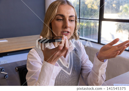 Female professional in business attire speaking into smartphone at desk in office Female professional in business attire speaking into smartphone at desk in office 141545375