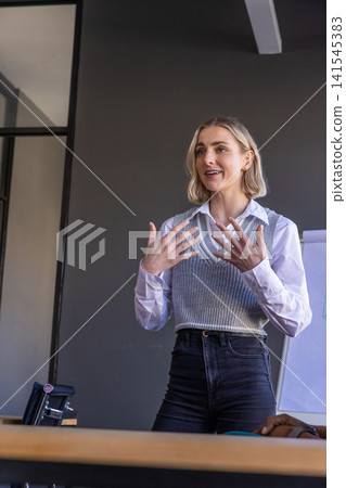 Female business professional presenting and gesturing in meeting room near flip chart, briefcase Female business professional presenting and gesturing in meeting room near flip chart, briefcase 141545383