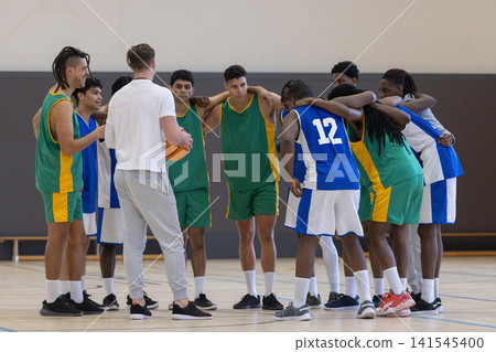 Diverse male basketball team huddling around coach holding basketball on polished wood court 141545400