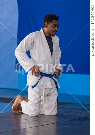 African american man kneeling on dojo mat adjusting blue belt, white gi, copy space African american man kneeling on dojo mat adjusting blue belt, white gi, copy space 141545403