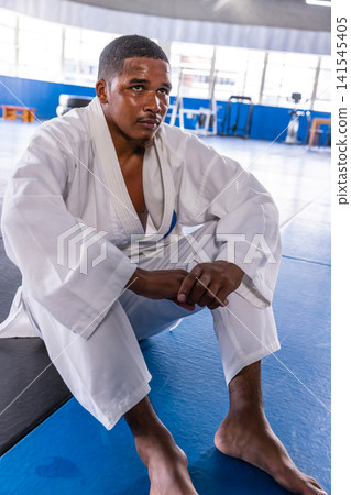 African american male martial artist sitting on blue mats in dojo amid kettlebells in white gi African american male martial artist sitting on blue mats in dojo amid kettlebells in white gi 141545405