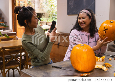 Asian daughter and senior mother carving pumpkins and photographing with phone at home dining table Asian daughter and senior mother carving pumpkins and photographing with phone at home dining table 141545423