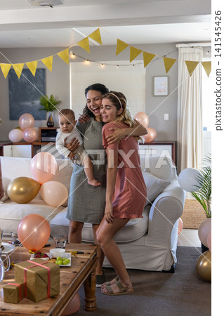 Diverse family hugging and celebrating in living room with gold pink balloons, gift boxes Diverse family hugging and celebrating in living room with gold pink balloons, gift boxes 141545426