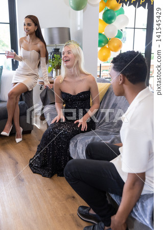 Diverse friends laughing and celebrating birthday in living room, balloons, tiara Diverse friends laughing and celebrating birthday in living room, balloons, tiara 141545429
