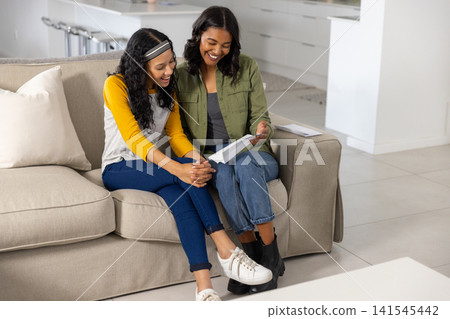 Diverse mom and teenage daughter reading letter from envelope while smiling on sofa at home Diverse mom and teenage daughter reading letter from envelope while smiling on sofa at home 141545442