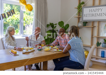 Diverse friends sitting around wooden dining table celebrating retirement with juice, cupcakes Diverse friends sitting around wooden dining table celebrating retirement with juice, cupcakes 141545444