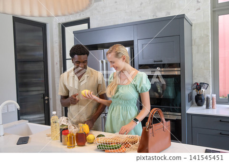 Diverse couple placing lemons and peppers into reusable bag with smartphone on kitchen countertop 141545451
