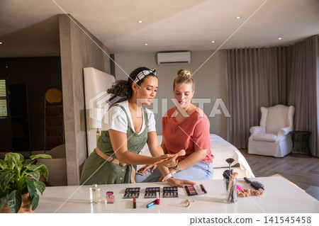 Diverse female friends demonstrating makeup swatches at bedroom tabletop with palette, brushes Diverse female friends demonstrating makeup swatches at bedroom tabletop with palette, brushes 141545458