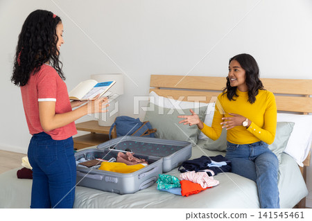 Diverse mom and teenage daughter packing grey suitcase with guidebook on bed in bedroom Diverse mom and teenage daughter packing grey suitcase with guidebook on bed in bedroom 141545461