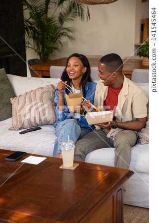 African American couple laughing and eating takeout with chopsticks on couch in living room African American couple laughing and eating takeout with chopsticks on couch in living room 141545464