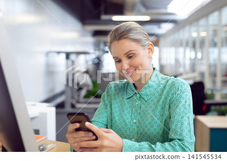 Adult woman wearing teal blouse sitting at desk in open office, holding smartphone near monitor Adult woman wearing teal blouse sitting at desk in open office, holding smartphone near monitor 141545534