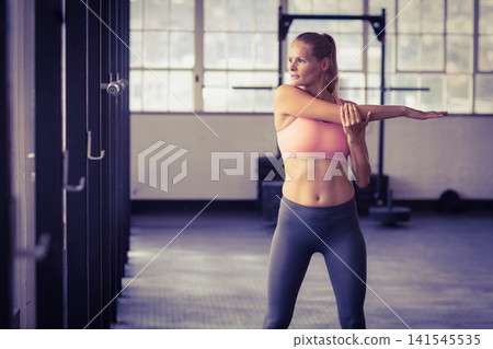 Female wearing pink bra and leggings doing shoulder stretch by squat rack in gym, copy space Female wearing pink bra and leggings doing shoulder stretch by squat rack in gym, copy space 141545535