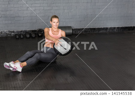 Female exerciser holding medball balancing with feet raised at gym wearing pink-bra by kettlebells Female exerciser holding medball balancing with feet raised at gym wearing pink-bra by kettlebells 141545538