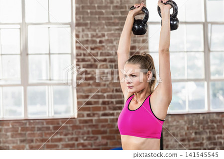 Female lifting kettlebells in bright pink sports bra by brick wall and multi-pane windows, blue mat Female lifting kettlebells in bright pink sports bra by brick wall and multi-pane windows, blue mat 141545545