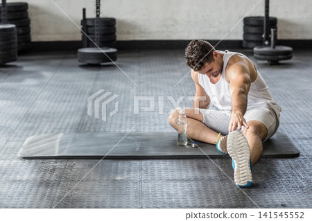 Man in 20s in tank stretching on gym mat, reaching toes to bottle, plates, copy space Man in 20s in tank stretching on gym mat, reaching toes to bottle, plates, copy space 141545552