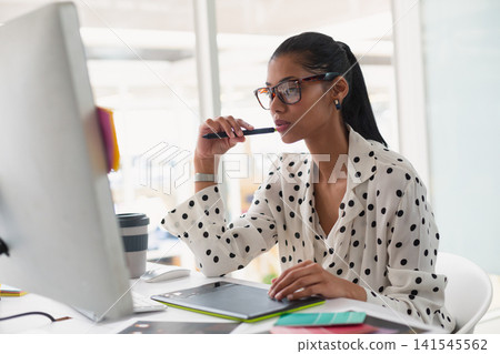 African American woman wearing polka-dot blouse sitting at desk holding stylus on graphics tablet African American woman wearing polka-dot blouse sitting at desk holding stylus on graphics tablet 141545562