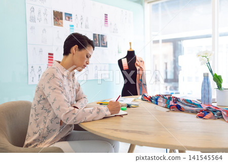 Mid-adult woman in pale pink blouse sketching at table in fashion studio with notebook and swatches Mid-adult woman in pale pink blouse sketching at table in fashion studio with notebook and swatches 141545564