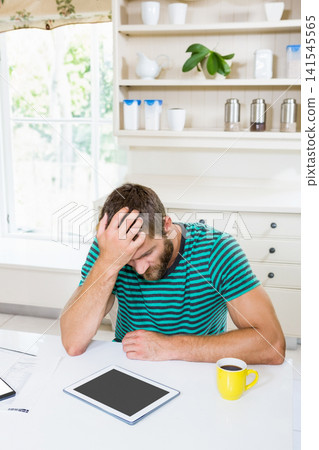 30s man wearing striped tee, leaning over tablet at kitchen table with yellow mug, papers 30s man wearing striped tee, leaning over tablet at kitchen table with yellow mug, papers 141545565