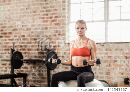 Female in orange sports bra and black leggings exercising on stability ball holding hex dumbbells Female in orange sports bra and black leggings exercising on stability ball holding hex dumbbells 141545567