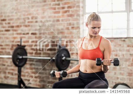 Woman wearing orange bra, black pants sitting on bench at gym holding hex dumbbells, copy space Woman wearing orange bra, black pants sitting on bench at gym holding hex dumbbells, copy space 141545568