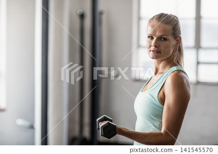 Female wearing light teal sleeveless top standing in gym, holding hex dumbbell by racks, copy space Female wearing light teal sleeveless top standing in gym, holding hex dumbbell by racks, copy space 141545570