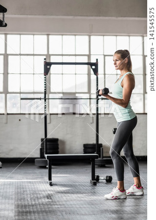 Woman in 30s, mint tank top lifting hex dumbbell at gym with squat rack, copy space Woman in 30s, mint tank top lifting hex dumbbell at gym with squat rack, copy space 141545575