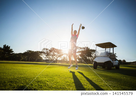 Female in late twenties jumping on golf fairway in pink polo, holding driver, white cart visible Female in late twenties jumping on golf fairway in pink polo, holding driver, white cart visible 141545579