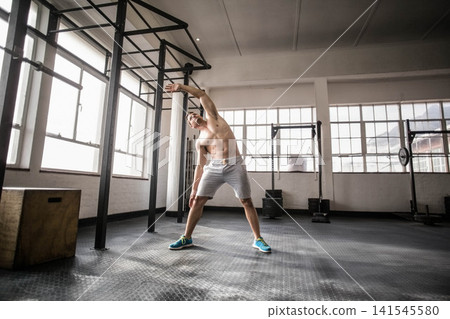 Adult male athlete performing side stretch at gym, pull-up rig, shirtless wearing shorts and shoes Adult male athlete performing side stretch at gym, pull-up rig, shirtless wearing shorts and shoes 141545580