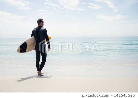 Man standing on sandy beach holding surfboard yellow nose in black wetsuit facing ocean, copy space Man standing on sandy beach holding surfboard yellow nose in black wetsuit facing ocean, copy space 141545586