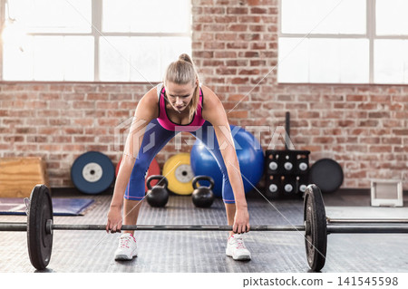 Adult female athlete wearing pink tank top and blue leggings, bending over loaded barbell at gym Adult female athlete wearing pink tank top and blue leggings, bending over loaded barbell at gym 141545598