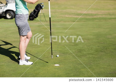 Adult female golfer standing holding flagstick near hole with golf ball, bag and cart, copy space Adult female golfer standing holding flagstick near hole with golf ball, bag and cart, copy space 141545600